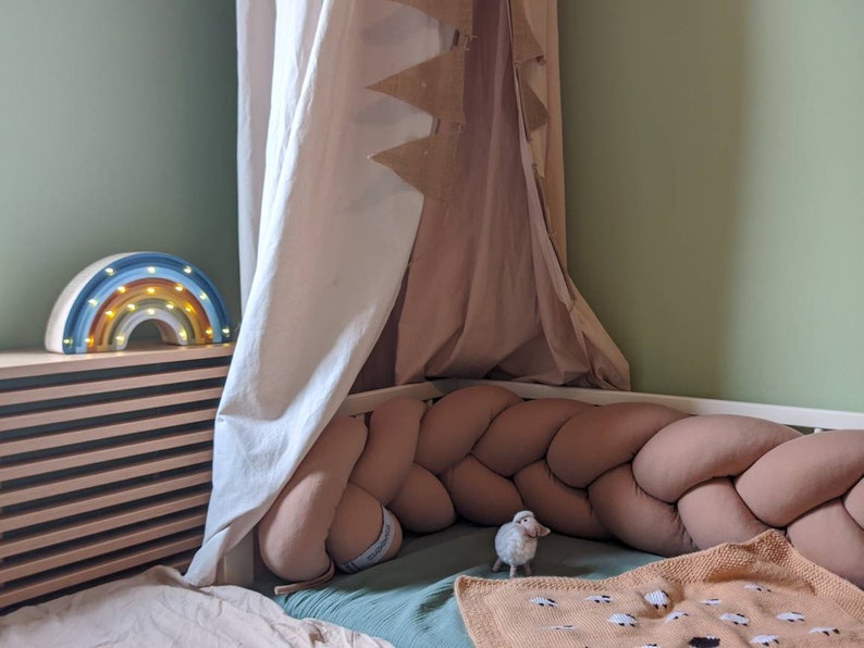May include: A children's bedroom scene featuring a rainbow-shaped night light with warm white lights. A braided bumper surrounds the bed, and a small sheep figurine sits near a blanket with sheep patterns. A canopy hangs above the bed.