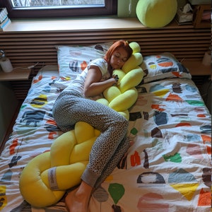 May include: A yellow braided pillow is on a bed with a patterned comforter. A person is sleeping on the bed, hugging the pillow. A large, round, yellow pillow is on the wall behind the bed.