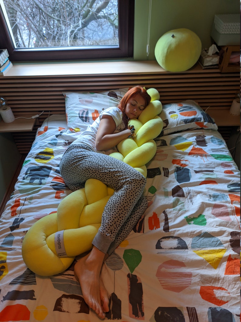 May include: A yellow braided pillow is on a bed with a patterned comforter. A person is sleeping on the bed, hugging the pillow. A large, round, yellow pillow is on the wall behind the bed.