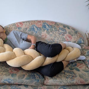 May include: A large, braided decorative pillow in a soft, light yellow color. The pillow is resting on a floral patterned sofa. The sofa has a vintage look with a mix of blue, pink, and green floral designs.