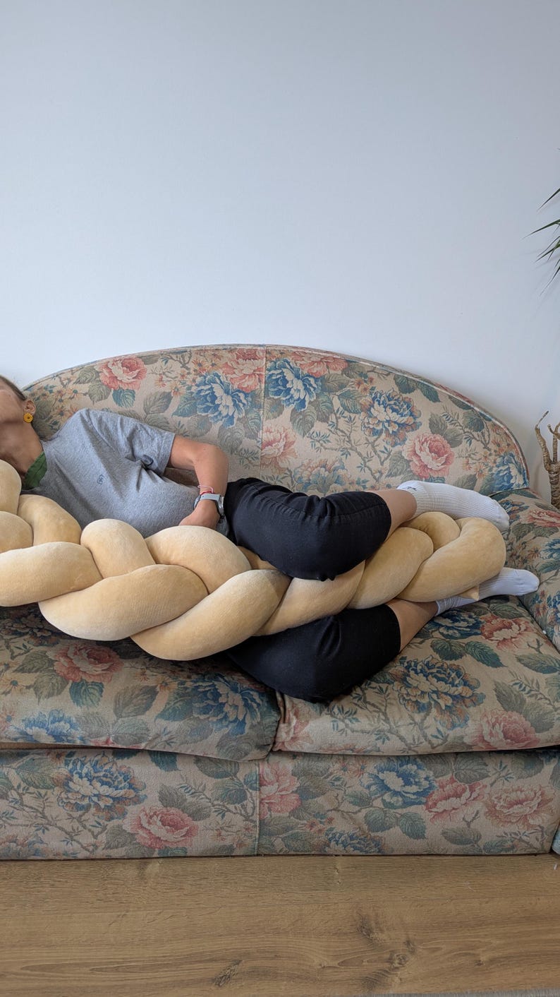 May include: A large, braided decorative pillow in a soft, light yellow color. The pillow is resting on a floral patterned sofa. The sofa has a vintage look with a mix of blue, pink, and green floral designs.
