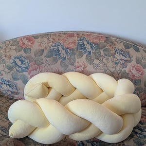 May include: A light yellow braided pillow, resting on a floral patterned sofa. The pillow is made of a soft, plush material and has a thick, woven design. The sofa has a vintage look with a mix of blue, pink, and green floral patterns.