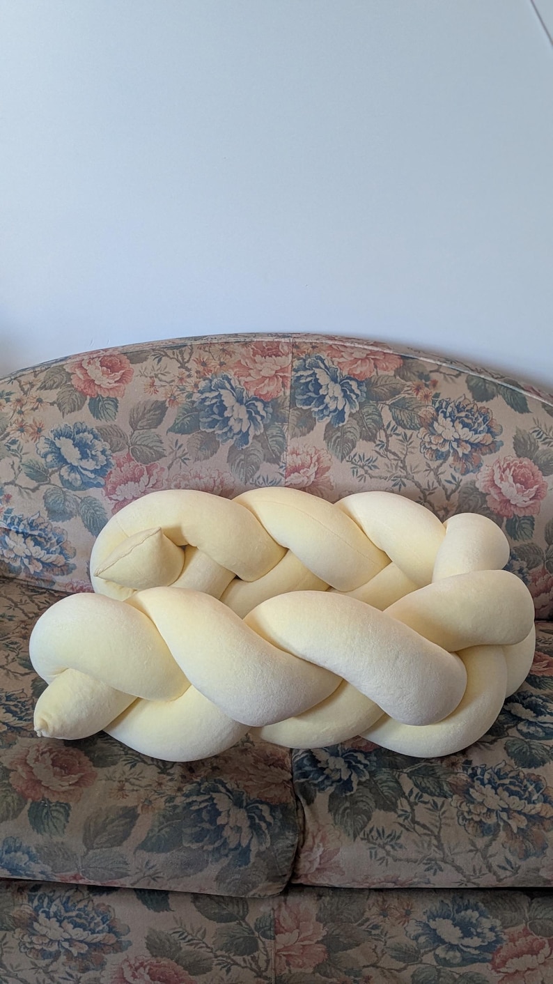 May include: A light yellow braided pillow, resting on a floral patterned sofa. The pillow is made of a soft, plush material and has a thick, woven design. The sofa has a vintage look with a mix of blue, pink, and green floral patterns.