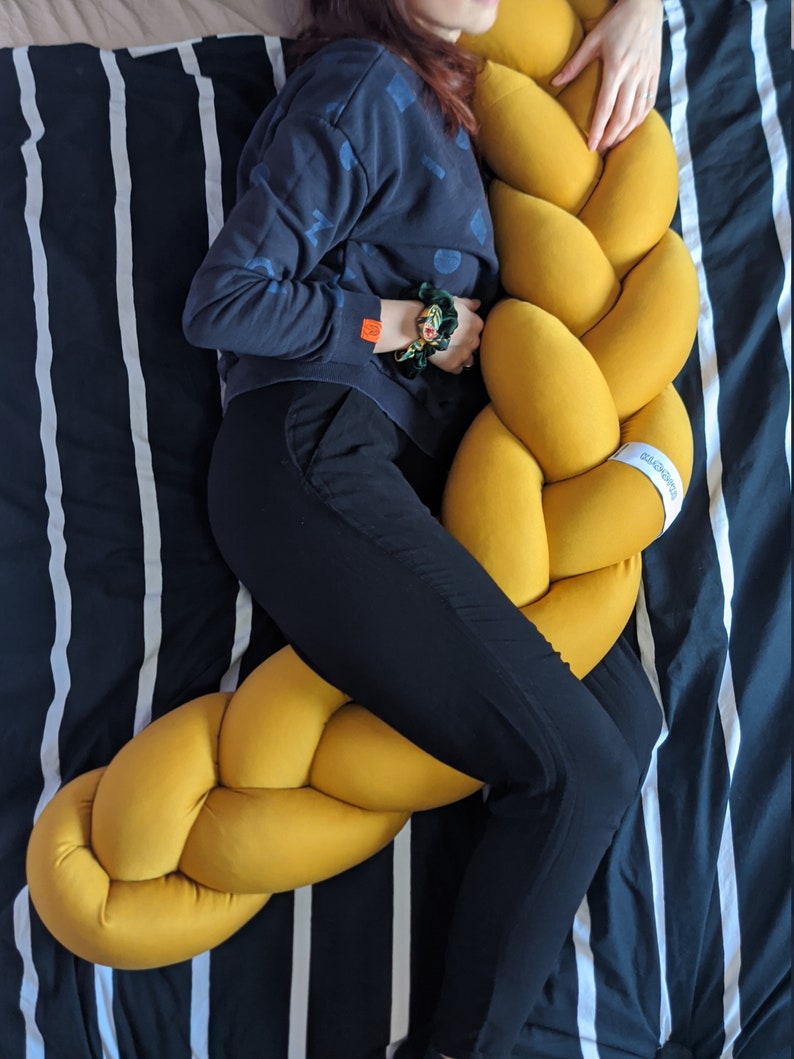 May include: A mustard yellow braided pillow rests on a bed with a black and white striped comforter. The pillow is a large, soft, and decorative item. The pillow is a unique home decor item.