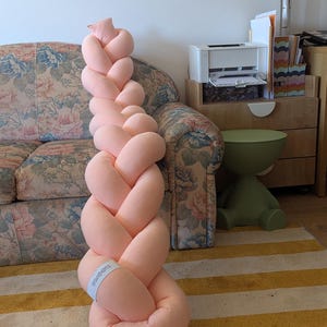 May include: A large, soft, braided pillow in a light peach color. The pillow is made of a plush material and is designed to resemble a long braid. It is placed on a striped rug with yellow and white stripes.