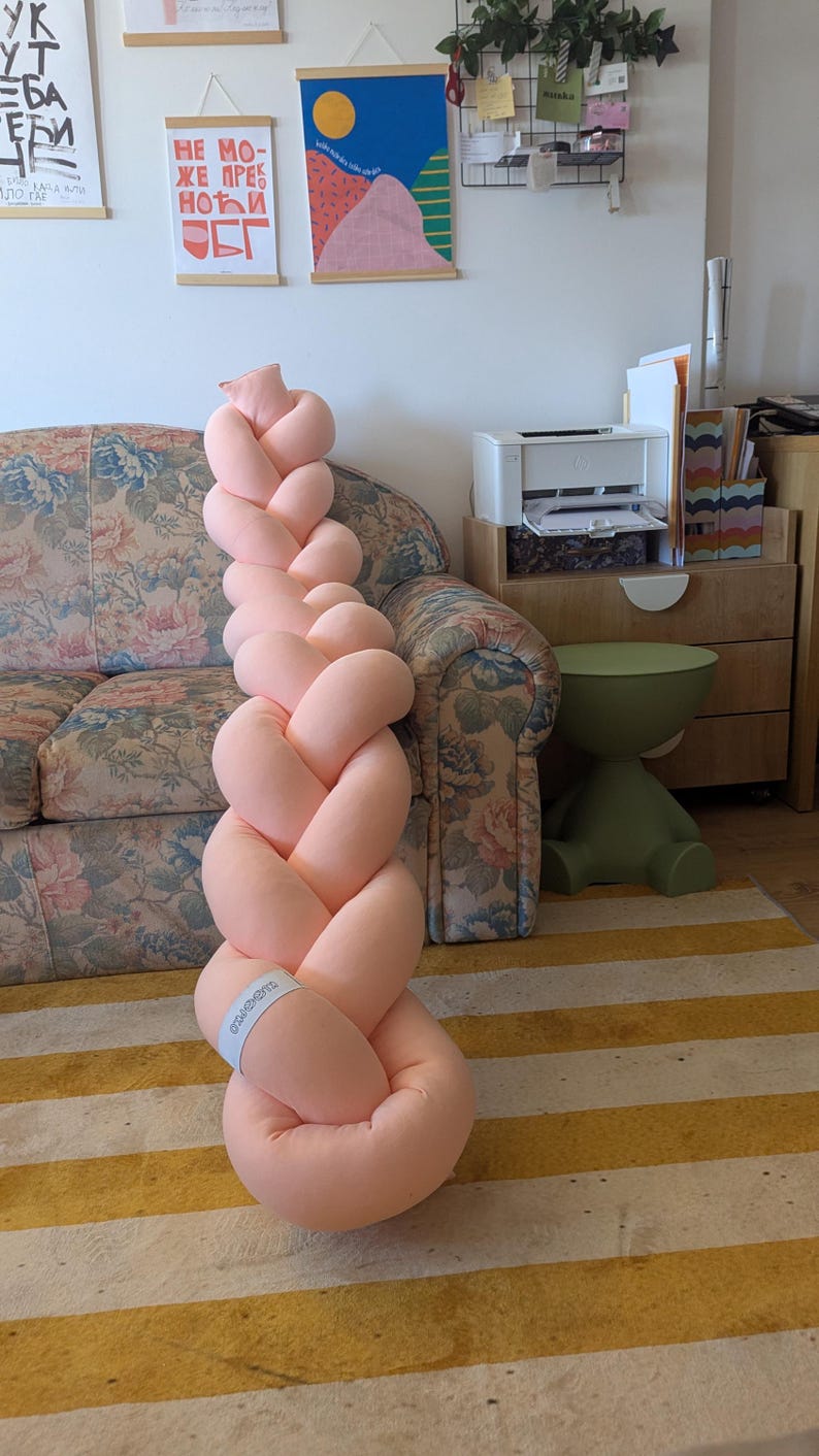 May include: A large, soft, braided pillow in a light peach color. The pillow is made of a plush material and is designed to resemble a long braid. It is placed on a striped rug with yellow and white stripes.