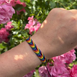 Könnte beinhalten: Ein gewebtes Freundschaftsarmband mit einem sich wiederholenden Fischgrätenmuster in leuchtenden Regenbogenfarben, darunter Rot, Orange, Gelb, Grün, Blau und Violett. Das Armband besteht aus dünnem Garn und hat einen einfachen schwarzen Kordelverschluss.
