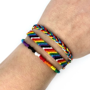 Peut inclure: Trois bracelets d'amitié colorés sur un poignet. Les bracelets sont faits de ficelle tissée en un motif arc-en-ciel, un motif rayé noir et blanc et un motif tressé avec de la ficelle rouge, jaune, bleue et noire.