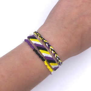 Puede incluir: Un conjunto de cuatro pulseras de la amistad en amarillo, morado, negro y blanco. Las pulseras están tejidas en una variedad de patrones, incluyendo chevron y trenza.