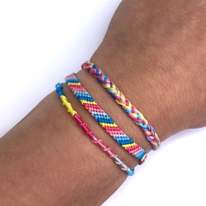 May include: Four colourful friendship bracelets on a wrist. The bracelets are made of woven string in shades of pink, blue, yellow, and grey.