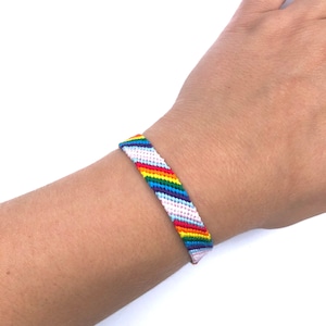 Pode incluir: Um bracelete de amizade arco-íris com listras brancas e rosa. O bracelete é feito de cordão tecido e é usado no pulso.
