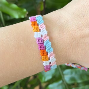 Peut inclure: Deux bracelets d'amitié tissés colorés. Les bracelets présentent un motif de fils roses, oranges, violets, blancs et bleus. Les bracelets sont portés au poignet, avec un feuillage vert en arrière-plan.