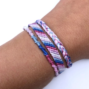 May include: Four friendship bracelets in pink, blue, and white colors. The bracelets are woven with a variety of patterns, including stripes and braids.