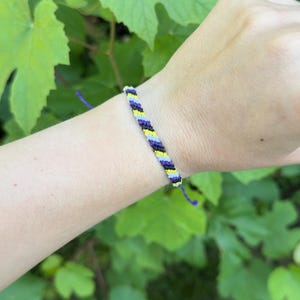 May include: A woven friendship bracelet featuring a repeating diagonal pattern of yellow, white, black, and purple thread. The bracelet is constructed from thin cord and includes adjustable tie ends that extend from the main braided section.