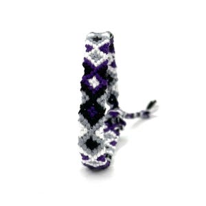 Peut inclure: Bracelet d'amitié fait à la main avec un motif géométrique noir, blanc, gris et violet. Le bracelet est fait de fil tissé et possède une fermeture nouée. Le motif comprend des formes de diamants et d'étoiles.