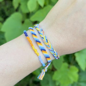 May include: Three handmade friendship bracelets featuring a color palette of bright yellow, orange, light blue, and dark blue. The set includes one braided bracelet and two thinner knotted bracelets with intricate woven patterns and loose ends.