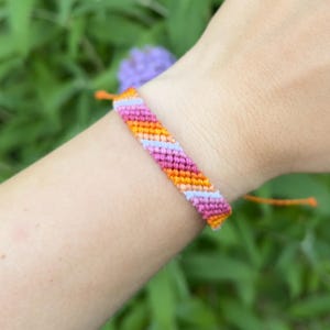 以下が含まれることがあります： オレンジ、ピンク、ライトパープルの斜めストライプ模様が特徴の織り込み式フレンドシップブレスレット。細い糸で構成されており、織り込まれた部分の両端からオレンジ色の紐が伸びています。