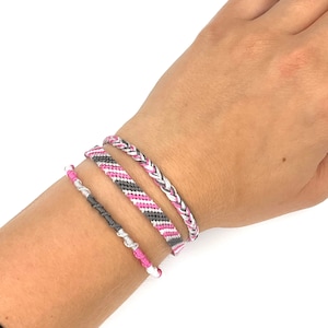 May include: Three friendship bracelets in pink, gray, and white. The bracelets are woven with a variety of patterns, including a braided pattern and a striped pattern.