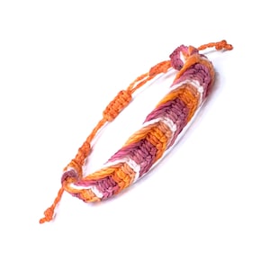 Puede incluir: Pulsera de macramé hecha a mano en tonos naranja, rosa y blanco. La pulsera tejida presenta un cierre de nudo corredizo ajustable. Un accesorio elegante para el uso diario.
