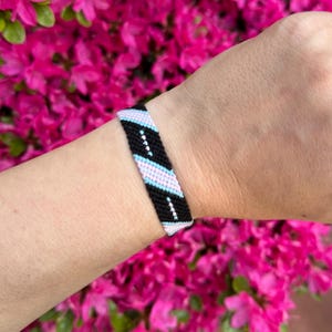 May include: A woven friendship bracelet in black, light blue, and white, with a diagonal pattern. The bracelet is worn on a wrist, set against a backdrop of vibrant pink flowers.
