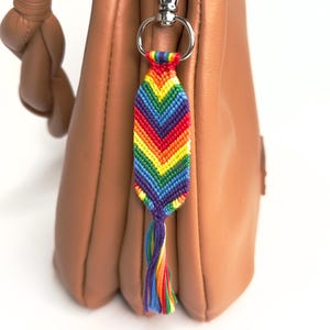 Può includere: Un portachiavi intrecciato con motivo a chevron arcobaleno e una piccola nappa sul fondo, fissato a un moschettone in metallo argentato. Il portachiavi presenta fili vivaci rossi, arancioni, gialli, verdi, blu e viola disposti a punta.