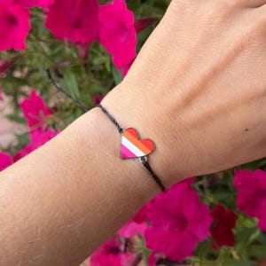 May include: A black cord bracelet featuring a small, heart-shaped metal charm. The charm is filled with enamel stripes in shades of orange, white, and pink, creating a horizontal pattern across the center of the heart-shaped pendant.