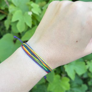 May include: A multi-strand cord bracelet featuring thin, vibrant threads in red, orange, yellow, green, blue, and purple. The bracelet is constructed from several individual colored strings arranged in a parallel pattern, secured with a simple knot closure.