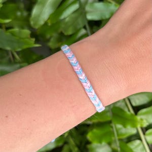 Könnte beinhalten: Geflochtenes Freundschaftsarmband in Pastellblau, Rosa und Weiß. Das Armband besteht aus gewebten Fäden, die ein strukturiertes Muster bilden. Das Armband wird am Handgelenk getragen.