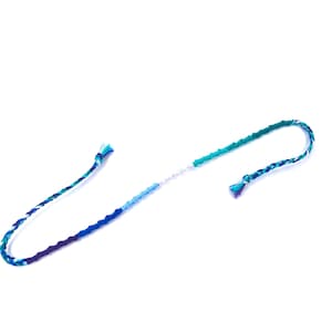 Puede incluir: Un brazalete de amistad multicolor con hilos azules, verdes y blancos. El brazalete est&aacute; atado en un nudo en ambos extremos.