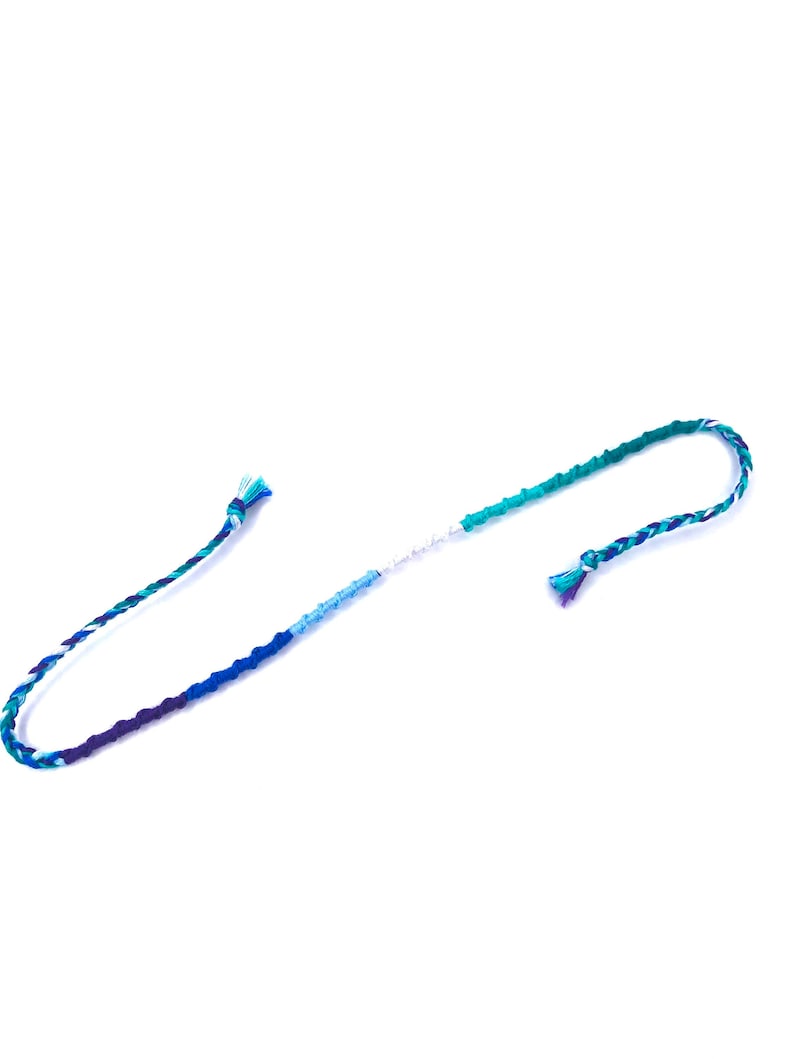Puede incluir: Un brazalete de amistad multicolor con hilos azules, verdes y blancos. El brazalete est&aacute; atado en un nudo en ambos extremos.