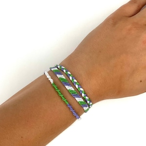 May include: Three friendship bracelets in green, white, and purple colors. The bracelets are woven together in a braided pattern.