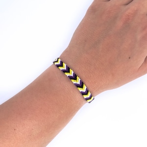 May include: A chevron patterned friendship bracelet. The bracelet is made of woven threads in white, yellow, black, and purple. The bracelet is worn on a wrist.