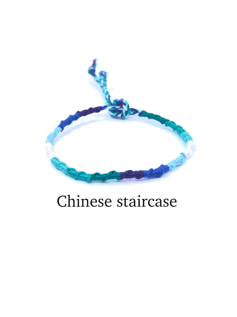 Puede incluir: Un brazalete de amistad trenzado azul, verde, morado y blanco con un nudo en el extremo. El brazalete se llama "escalera china".