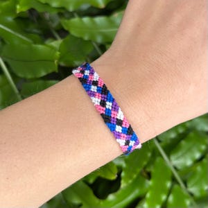 May include: A colorful friendship bracelet with a woven pattern. The bracelet features a repeating design of black, white, pink, blue, and purple threads. The bracelet is approximately 0.5 inches wide.