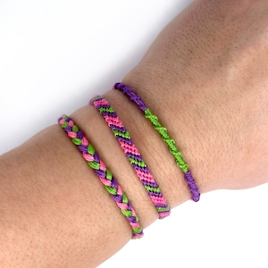 May include: Four colourful friendship bracelets. The bracelets are made of woven thread in shades of purple, pink, and green. One bracelet is braided, and another is twisted. The bracelets are displayed on a person's wrist.