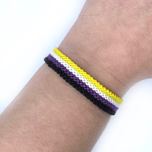 Pode incluir: Um bracelete de amizade roxo, branco e amarelo. O bracelete é tecido com um padrão simples e está amarrado no pulso de uma pessoa.