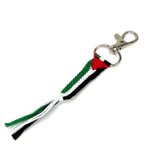 Puede incluir: Un llavero con un diseño tejido con los colores de la bandera palestina: rojo, blanco, verde y negro. El llavero tiene un cierre plateado y una anilla para sujetar las llaves. La sección tejida mide aproximadamente 10 cm de largo.