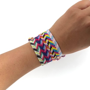 Könnte beinhalten: Eine Sammlung farbenfroher Freundschaftsarmbänder. Jedes Armband hat ein Zickzackmuster in verschiedenen Farben, darunter Rosa, Blau, Gelb und Schwarz. Die Armbänder sind an einem Handgelenk gestapelt und zeigen die lebendigen Designs.