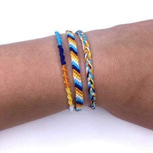 Può includere: Tre braccialetti dell'amicizia in blu, giallo e bianco. I braccialetti sono intrecciati con diversi motivi e vengono indossati al polso.