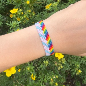 Op de afbeelding: Een regenboog vriendschapsarmband met roze, blauwe en witte strepen. De armband is geweven met een chevron-patroon.