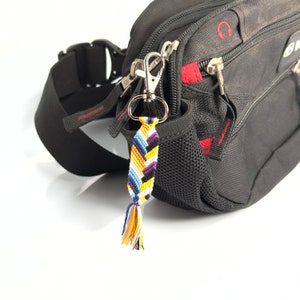 Puede incluir: Una riñonera negra con un bolsillo con cremallera y un bolsillo de malla. Un llavero tejido de colores con un borla cuelga del tirador de la cremallera.