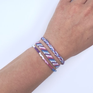 May include: A set of four friendship bracelets in shades of purple, pink, and white. The bracelets are woven with a chevron pattern and are tied together on a wrist.