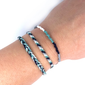 May include: Four braided friendship bracelets in shades of teal, white, and black. The bracelets are worn on a person's wrist.