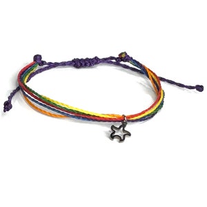 Può includere: Braccialetto dell'amicizia arcobaleno con un ciondolo a forma di stella. Il braccialetto presenta un design intrecciato con fili rossi, arancioni, gialli, verdi, blu e viola. Il ciondolo è di colore argento scuro e pende dal centro del braccialetto.
