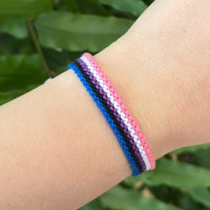 Peut inclure: Un bracelet d'amitié fait à la main avec un motif tissé de fils bleus, noirs, violets, blancs et roses. Le bracelet est porté au poignet, avec un feuillage vert en arrière-plan. Cet accessoire est un symbole de solidarité.
