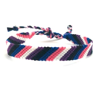 May include: A woven friendship bracelet with a pattern of black, blue, pink, white, and purple stripes.