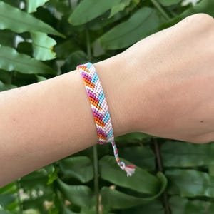 Peut inclure: Un bracelet d'amitié coloré avec un motif en chevrons. Le bracelet est composé de fils roses, oranges, bleus et blancs. Le bracelet est porté sur un poignet, avec des feuilles vertes en arrière-plan.