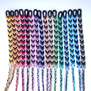 May include: A collection of 15 colourful chevron friendship bracelets with black accents. The bracelets are made with a variety of colours, including orange, pink, purple, green, blue, and grey.