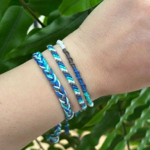 Op de afbeelding: Vier vriendschapsarmbanden in de kleuren blauw, wit en grijs. De armbanden zijn geweven met verschillende patronen, waaronder een gevlochten stijl.