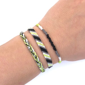 Puede incluir: Tres pulseras de la amistad en colores negro, blanco y verde. Las pulseras están tejidas con un patrón simple y están atadas en la muñeca de una persona.